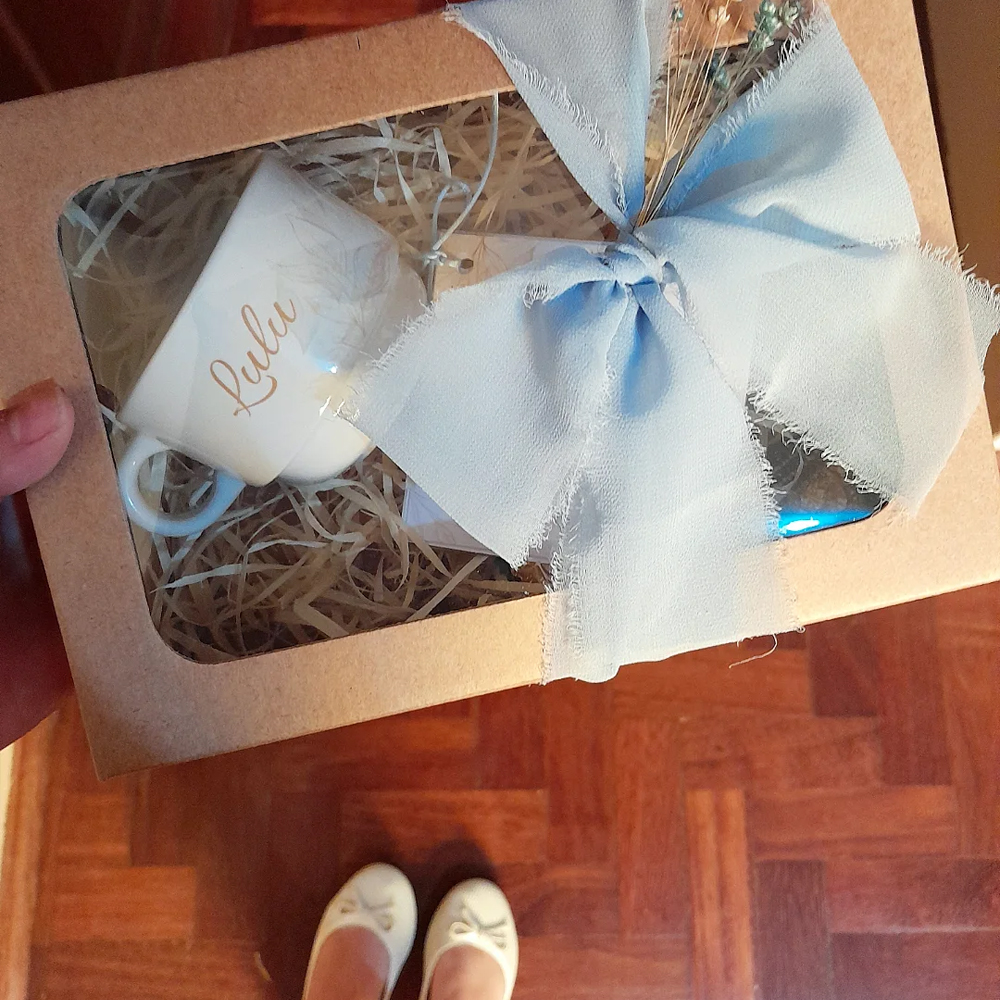 Caixa de presente de papel kraft com tampa de acetato transparente, vista de cima. Dentro, há palha decorativa e uma xícara branca com o nome Lulu escrito em dourado. A caixa está decorada com um laço grande de tecido azul-bebê. A foto é tirada de cima, mostrando também os pés da pessoa que segura a caixa: ela usa sapatilhas branca com detalhe de lacinho. O chão é de madeira.