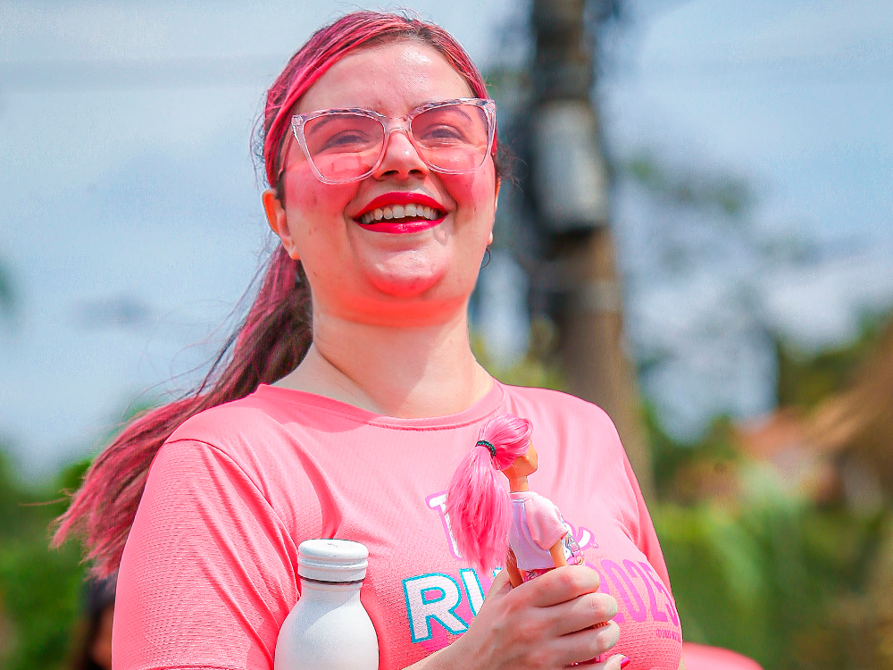 Luly sorridente em close durante a corrida. Ela usa camiseta rosa, óculos transparentes com leve tom rosado e a cabelo, também rosa, está preso em um rabo de cavalo que se movimenta com o vento. Na mão direita, segura uma garrafinha branca e uma boneca Barbie de cabelo rosa. Ao fundo, árvores desfocadas e postes compõem um cenário externo iluminado pelo sol.