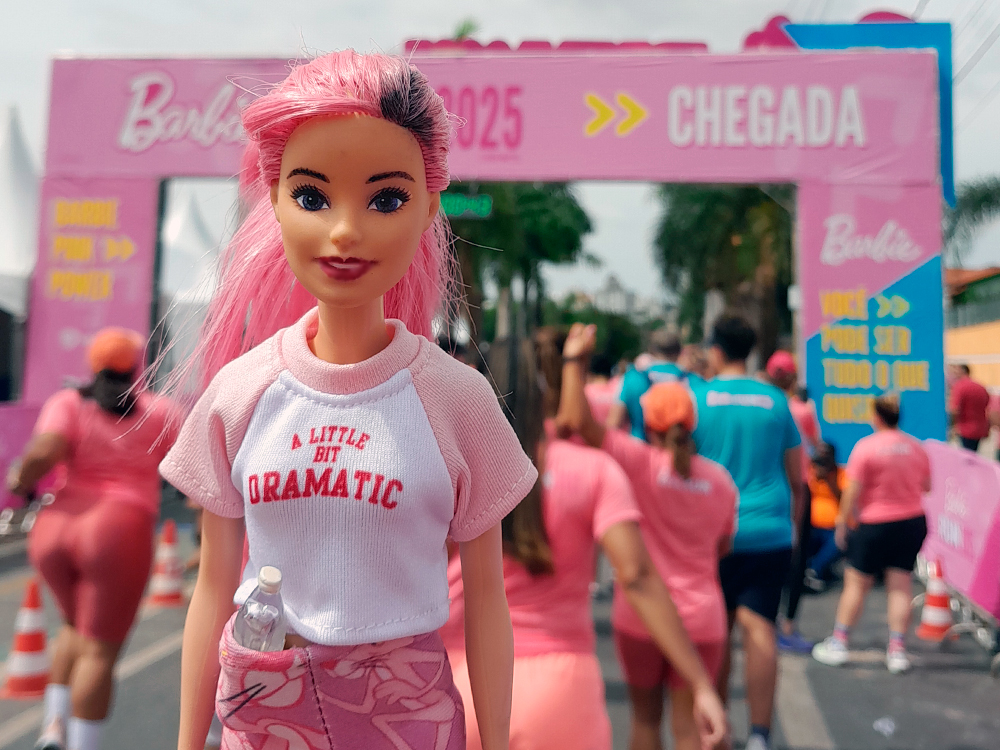 Boneca Barbie de cabelo rosa em destaque no primeiro plano, com uma camiseta branca e rosa escrita “A LITTLE BIT DRAMATIC”. A boneca segura uma mini garrafinha encaixada no cós da calça. Atrás dela, um grande portal de chegada da Barbie Run 2025 em tons de rosa e azul, com as palavras “CHEGADA” e setas amarelas. Participantes vestindo roupas esportivas também em rosa e azul caminham pela rua, cercada por cones laranja.