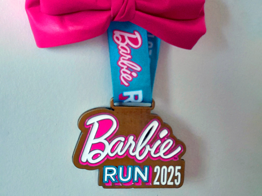 Close de uma medalha da BarbieRun 2025 pendurada por uma fita larga azul estampada com a palavra Barbie em rosa e RUN em branco. A medalha tem formato orgânico, lembrando uma nuvem ou placa estilizada, com fundo dourado metálico. No centro, o logo Barbie aparece em letras cursivas brancas com borda rosa vibrante. Logo abaixo, RUN 2025 está escrito em fonte sem serifa. A medalha está apoiada sobre um fundo claro, e no topo da fita há um laço grande e rosa-choque.