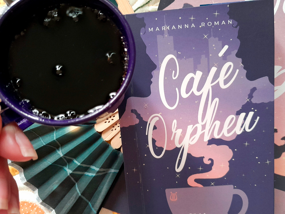Close de uma mão segurando uma caneca roxa cheia de café preto, ocupando o lado esquerdo da imagem. À direita, o livro Café Orpheus aparece em destaque. Ao fundo, aparece parte do leque azul e da superfície colorida onde a composição foi montada.