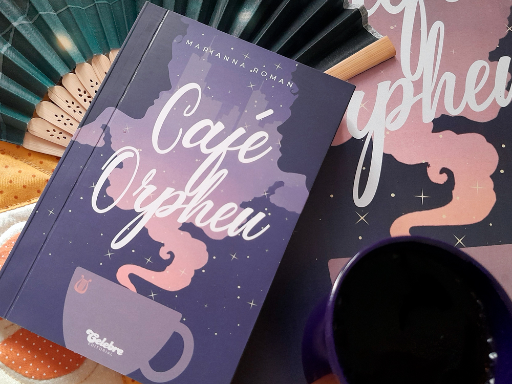 Flat lay do livro posicionado levemente inclinado sobre seu pôster com a mesma arte da capa. À esquerda, aparece o leque azul aberto; no canto inferior direito, parte da caneca roxa.