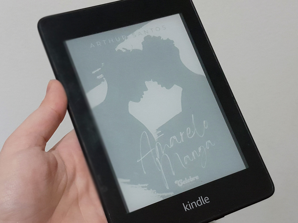 Mão esquerda segurando um Kindle preto contra um fundo neutro claro. Na tela, aparece a capa do livro Amarelo Manga, de Arthur Santos, em tons de cinza típicos do e-ink. A arte mostra a silhueta de duas pessoas prestes a se beijar, posicionadas de perfil, com os rostos se aproximando no centro da imagem. O título, em tipografia manuscrita, aparece na parte inferior da capa.