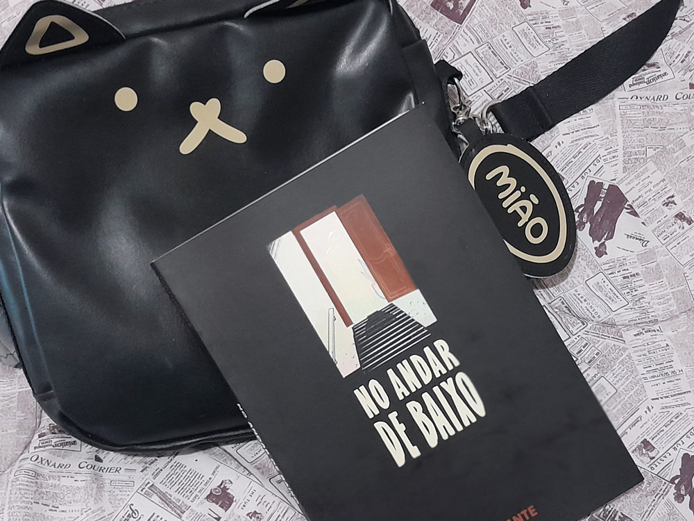 Livro No Andar de Baixo, com capa preta e ilustração de uma porta aberta no topo de uma escada, apoiado sobre uma bolsa preta em formato de gatinho. A bolsa é feita de material liso, com o rosto do gato estilizado em linhas claras (olhos circulares, focinho em “X” e orelhas triangulares). Ao lado, uma tag oval pendurada na alça mostra a palavra mião. O fundo é composto por folhas estampadas com páginas de jornal vintage.