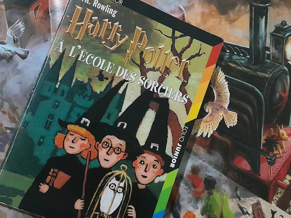 Edição francesa do livro Harry Potter à l’École des Sorciers, da coleção Folio Junior, posicionada sobre um fundo ilustrado com elementos do universo da série (como o Expresso de Hogwarts e corujas brancas em voo). A capa mostra uma ilustração estilizada dos três protagonistas usando chapéus pontudos, acompanhados de outros personagens em frente ao castelo de Hogwarts, tudo em traços simples e cores sóbrias.