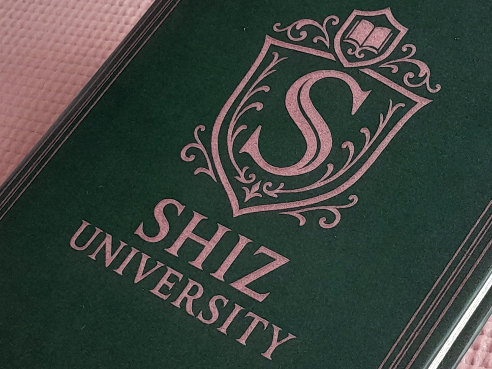 Caderno verde-escuro com capa dura e detalhes ornamentais em rosa metálico, exibindo o brasão da “Shiz University” ao centro. O fundo é uma superfície rosa texturizada.