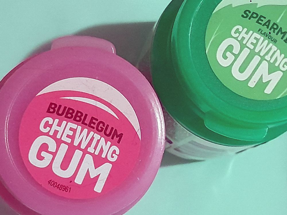 Vista superior de dois potes de chiclete, um rosa sabor bubblegum e outro verde sabor spearmint, sobre uma superfície verde-água suave.
