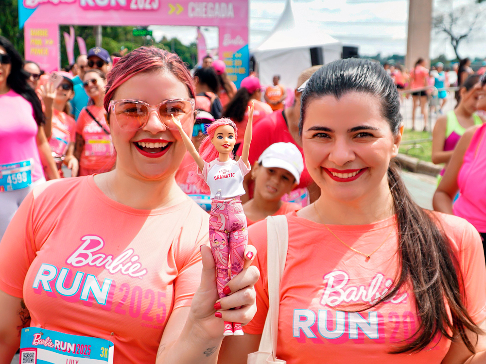 Luly e sua irmã sorriem para a câmera durante, as duas muito parecidas, mas uma de cabelos rosa e a outra castanha, segurando juntas uma Barbie de cabelo rosa vestida com camiseta branca e calça estampada. As duas usam camisetas temáticas da corrida, cercadas por outras participantes no clima animado do evento.