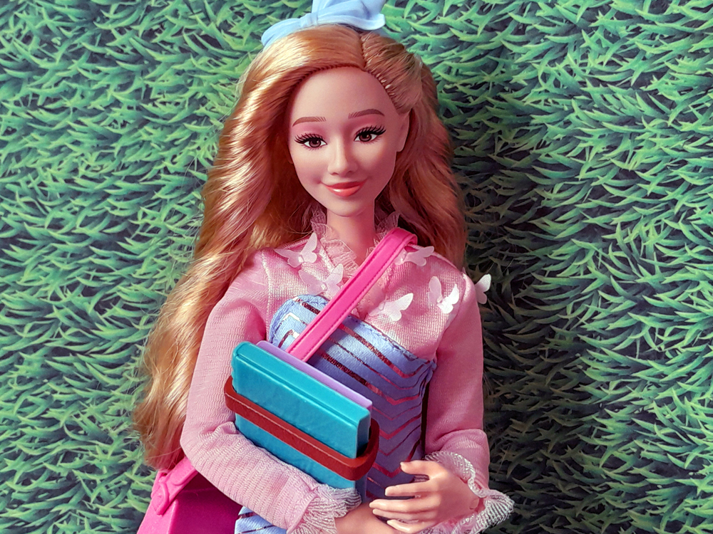 Boneca da Glinda de Wicked, da Mattel, em retrato frontal, com cabelo loiro ondulado, laço azul na cabeça e bolsa rosa atravessada no corpo, segurando livros coloridas amarrados por um cinto.