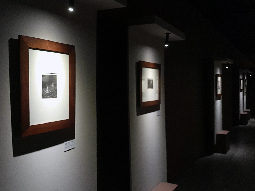 Corredor de exposição com gravuras de Rembrandt em molduras de madeira, alinhadas em painéis claros ao longo da parede, com iluminação direcionada e bancos baixos posicionados à frente das obras para contemplação.