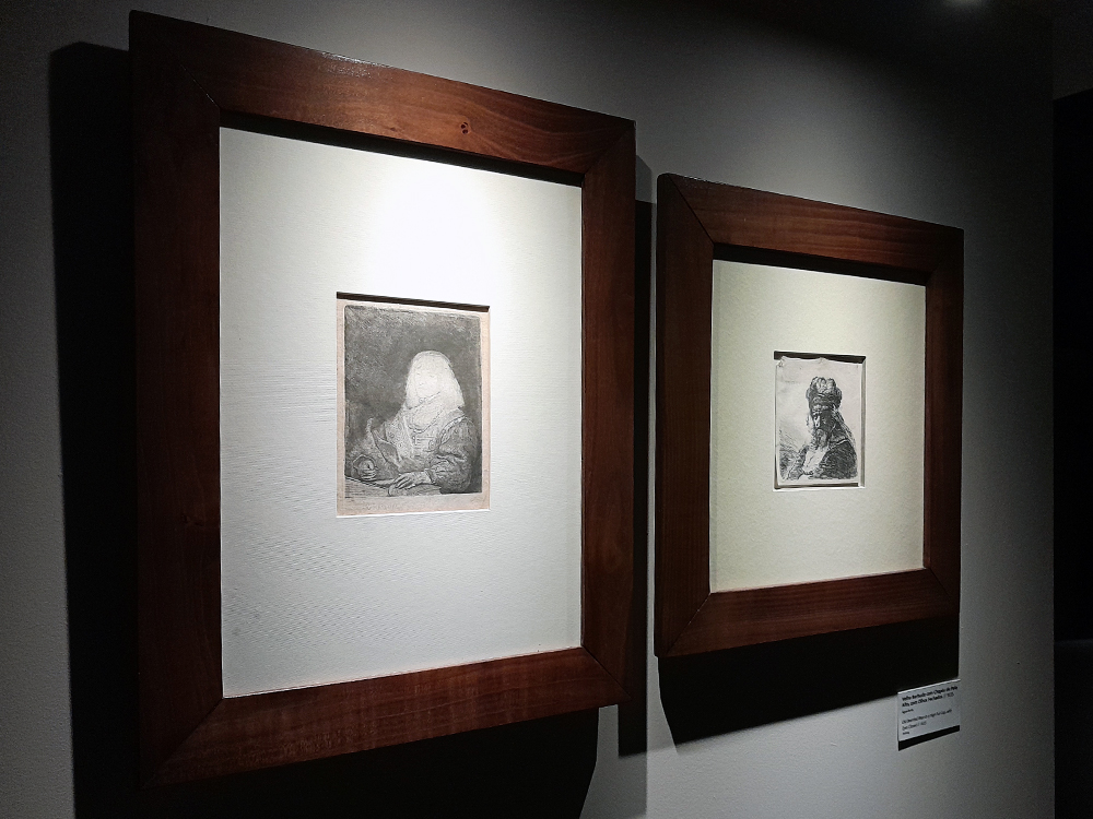 Duas gravuras expostas lado a lado, cada uma em moldura de madeira escura com paspatur claro, destacando retratos em preto e branco sob iluminação focada de museu.