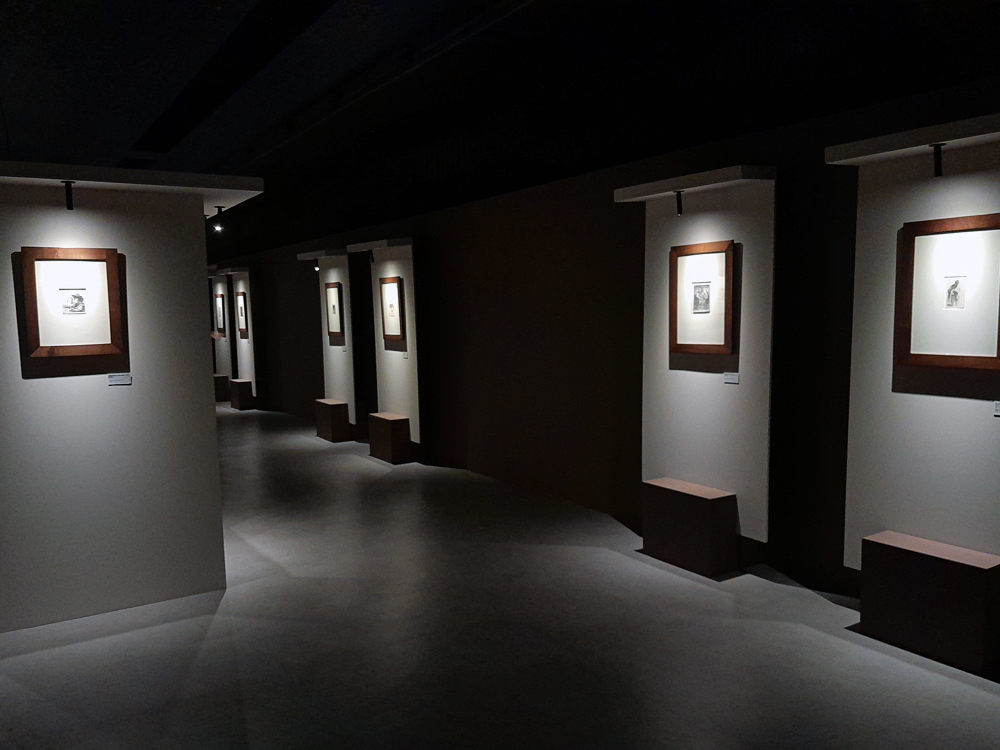 Vista ampla da exposição de Rembrandt, mostrando vários painéis expositivos ao longo de uma sala escura, com gravuras emolduradas iluminadas individualmente e um percurso contínuo que conduz o visitante pelo espaço.
