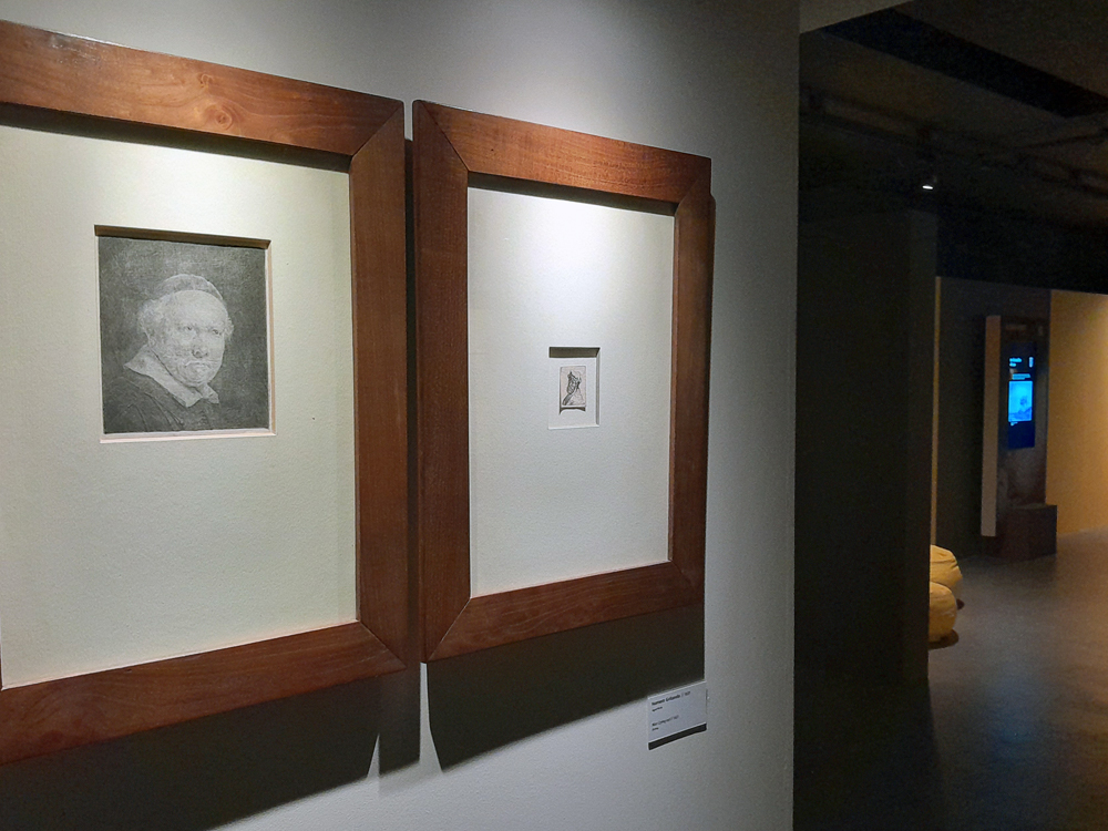 Gravura em preto e branco representando um retrato masculino, exibida em moldura de madeira escura com paspatur claro, sob iluminação suave em ambiente museológico.