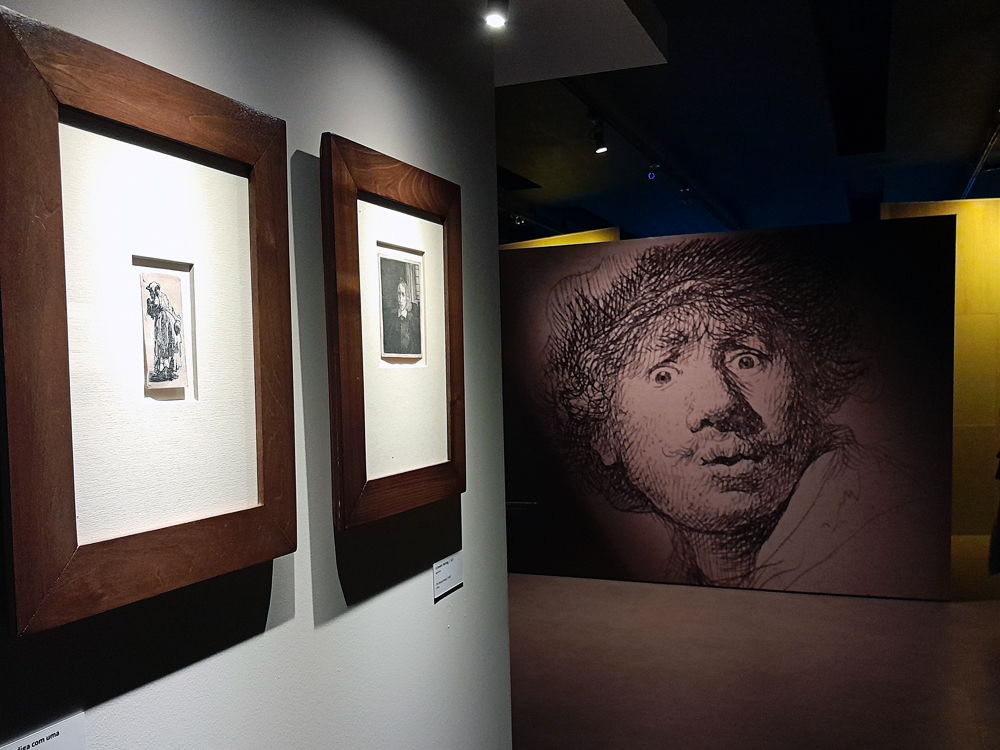 Vista da exposição de Rembrandt na Casa Fiat de Cultura, com gravuras emolduradas expostas na parede e, ao fundo, uma ampliação monumental de um autorretrato do artista.