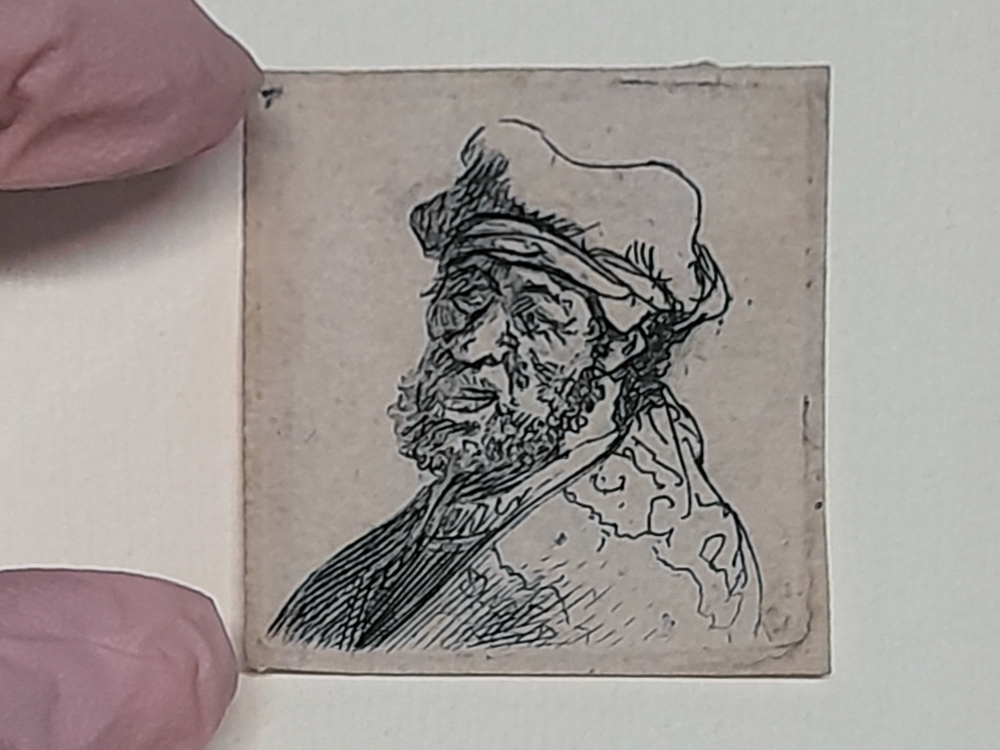 Gravura de Rembrandt de dimensões muito reduzidas, do tamanho aproximado de um selo, representando o retrato de um homem barbado usando chapéu; a obra é segurada por dedos cobertos com luvas rosa, indicando o manuseio técnico durante a elaboração de laudos de conservação e ajudando a dimensionar a escala diminuta da peça.