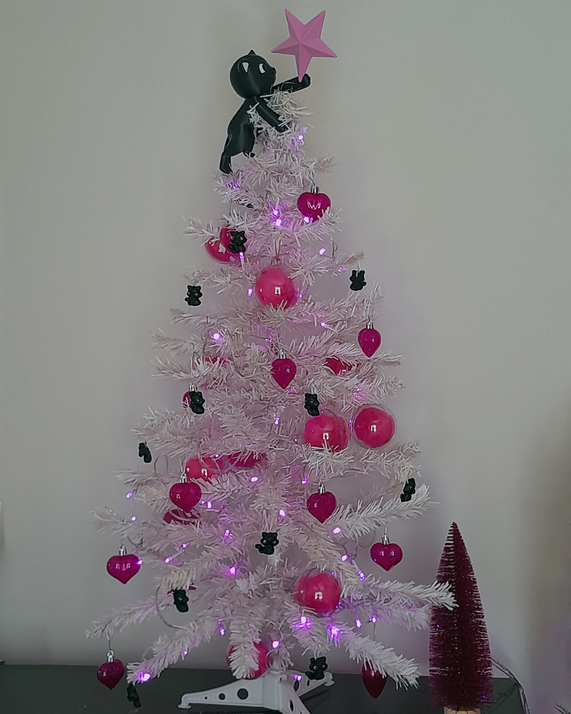 Árvore de Natal branca posicionada contra uma parede clara, decorada com luzes rosadas acesas e enfeites predominantemente rosa, incluindo bolas brilhantes e pingentes em formato de coração. Entre os galhos, pequenos ornamentos escuros em formato de gatinhos aparecem distribuídos pela árvore. No topo, há um enfeite de gato preto estilizado segurando uma estrela rosa, substituindo a tradicional estrela natalina. Ao lado direito da árvore, no chão, aparece um pequeno pinheiro decorativo rosa-choque.