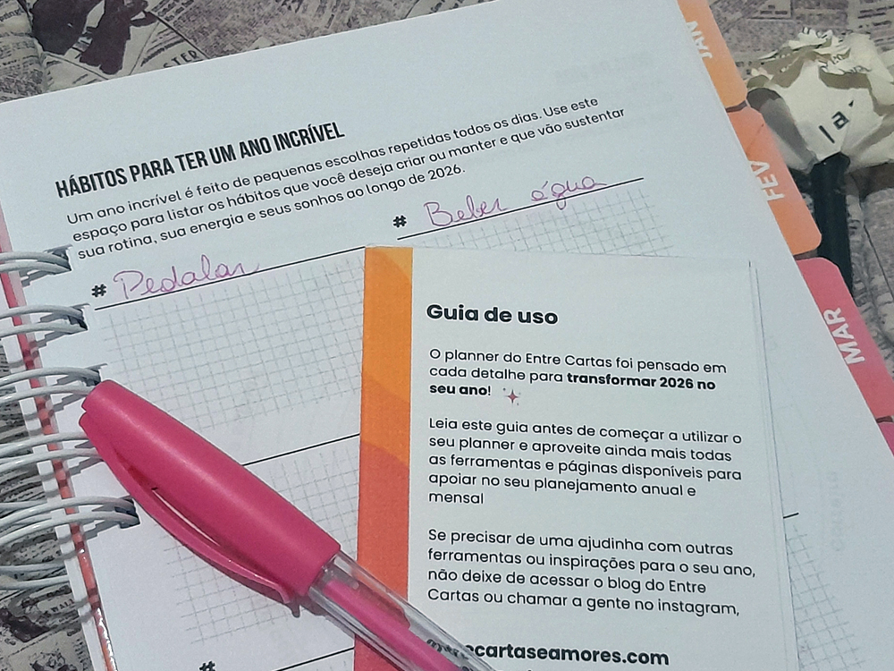 Páginas internas do planner do Entre Cartas e Amores mostrando a seção Hábitos para ter um ano incrível, com espaço para anotações como pedalar e beber água, além do guia de uso do planner, acompanhadas de uma caneta rosa e encadernação em espiral branca.