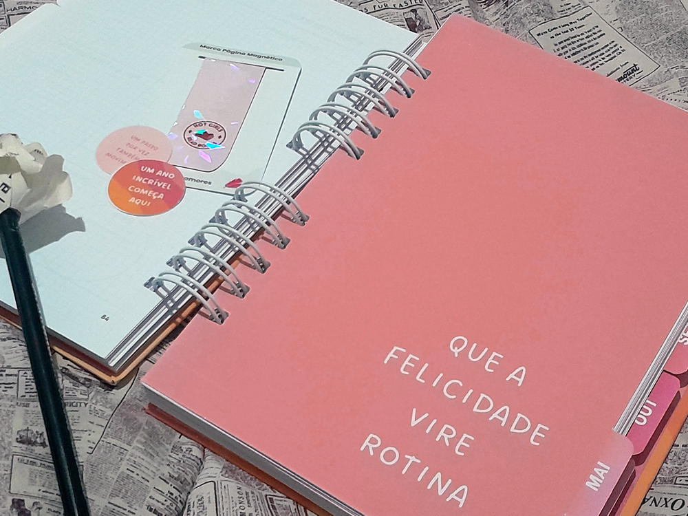 Divisória mensal referente a Maio em tom rosado, com a frase Que a felicidade vire rotina escrita em branco, ao lado de páginas internas abertas e detalhes da encadernação em espiral.