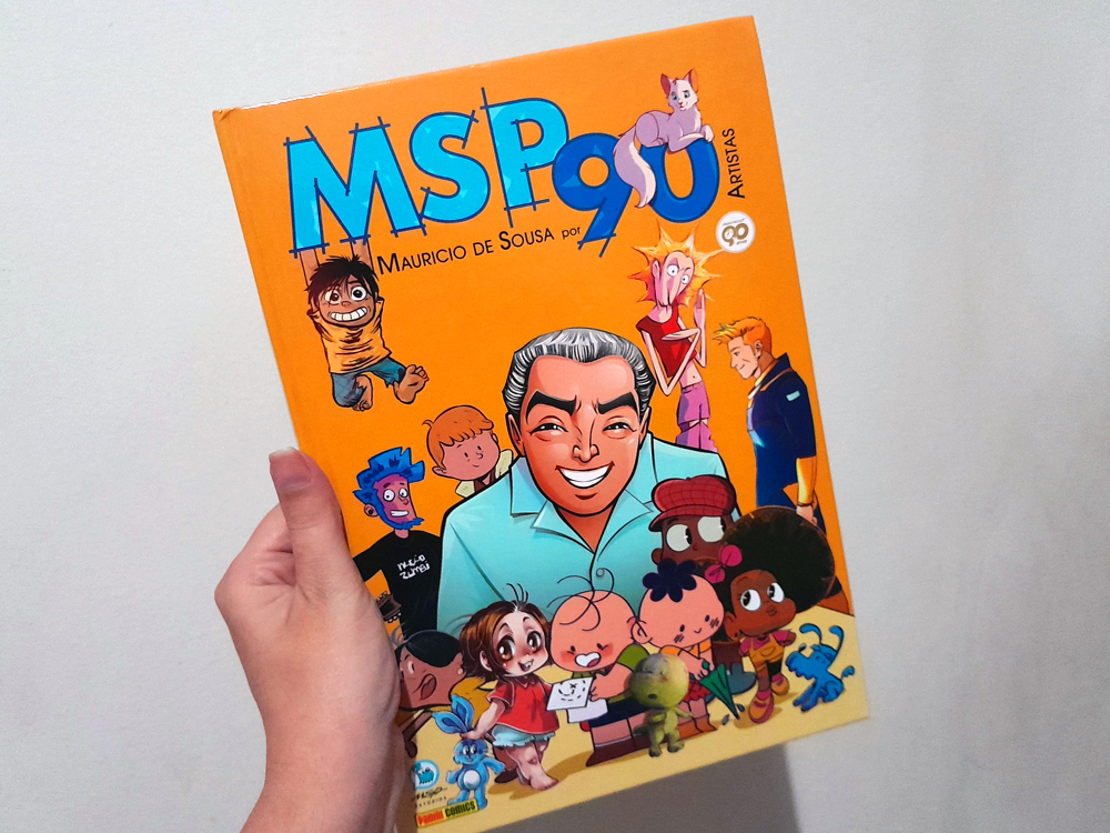 Mão segurando o livro MSP 90 – Mauricio de Sousa por artistas. A capa é amarela e traz uma ilustração de Mauricio de Sousa sorridente, cercado por vários personagens icônicos da Turma da Mônica e releituras feitas por diferentes artistas.