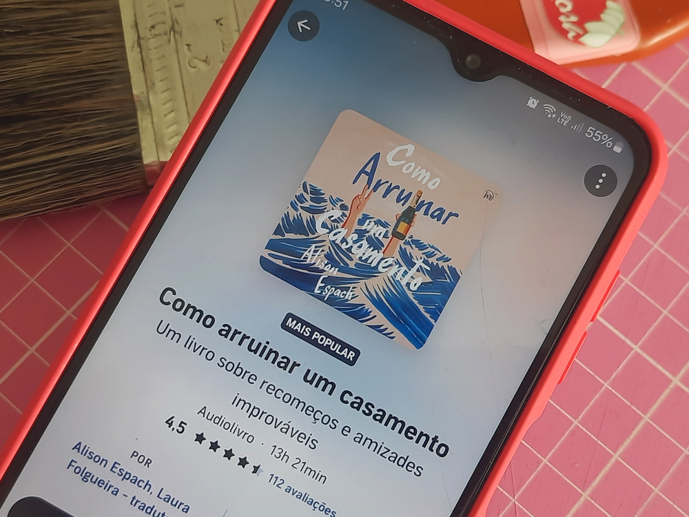 Smartphone com capa rosa exibindo a tela de um aplicativo de audiolivros. Na tela aparece o título Como arruinar um casamento, de Alison Espach, com a capa ilustrada em tons de azul e branco e informações como avaliação por estrelas e duração do áudio.