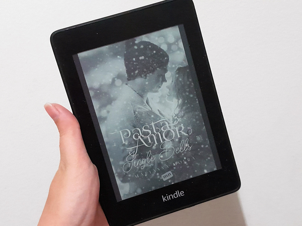 Mão segurando um Kindle para leituras om a capa do livro Pasta Amore & Single Bells, de Marianna Moura. A imagem da capa mostra um casal em clima romântico, em tons frios e acinzentados, com efeito de neve caindo ao fundo.