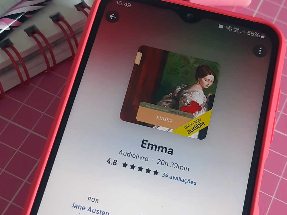 Smartphone com capa rosa exibindo a tela de um aplicativo de audiolivros. Na tela aparece o título Emma, de Jane Austen, com a capa verde contendo a imagem de uma mulher e informações como avaliação por estrelas e duração do áudio.