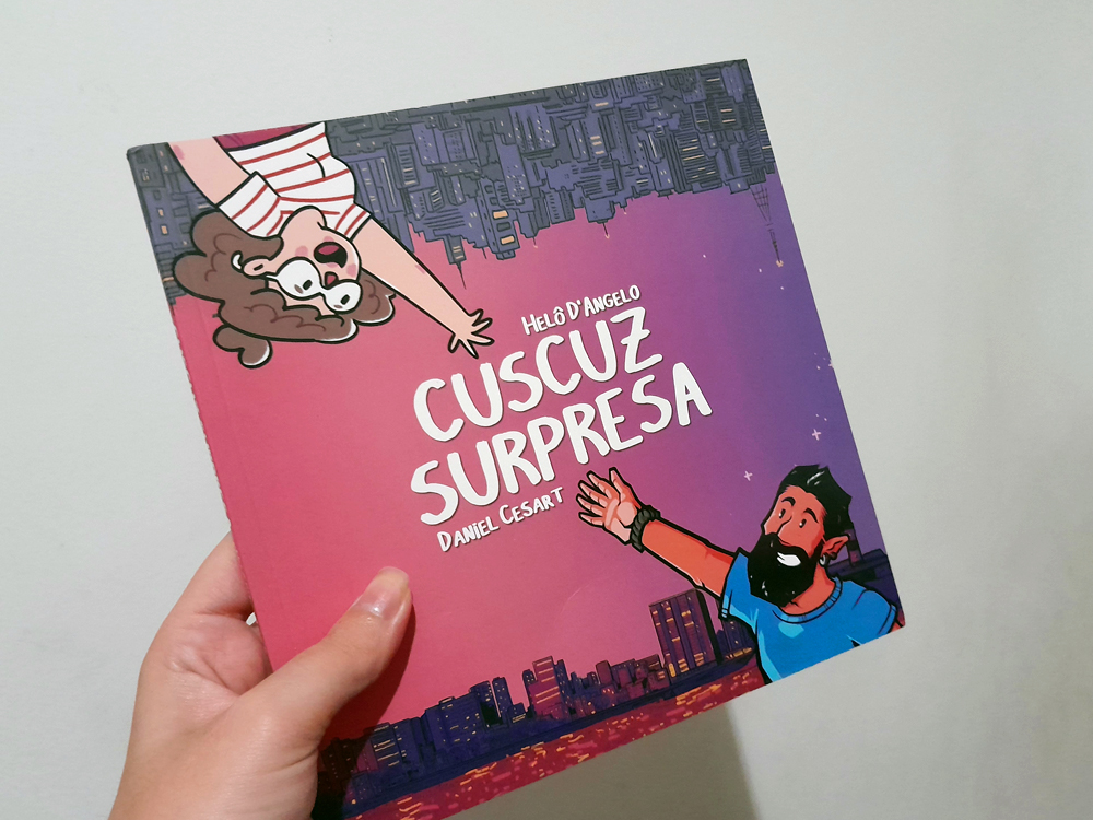 Mão segurando o livro Cuscuz Surpresa, de Helô D’Angelo e Daniel Cesar T. A capa ilustrada mostra dois personagens se estendendo um para o outro, com uma cidade ao fundo em tons de rosa, roxo e azul, criando um clima urbano e afetivo.