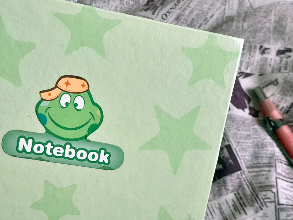 Caderno de capa verde-clara com estampa de estrelas em tom pastel e um desenho de sapinho sorridente usando chapéu, com a palavra Notebook em baixo. O caderno está apoiado sobre uma superfície com estampa de folhas de jornal, onde há uma lapiseira rosa.