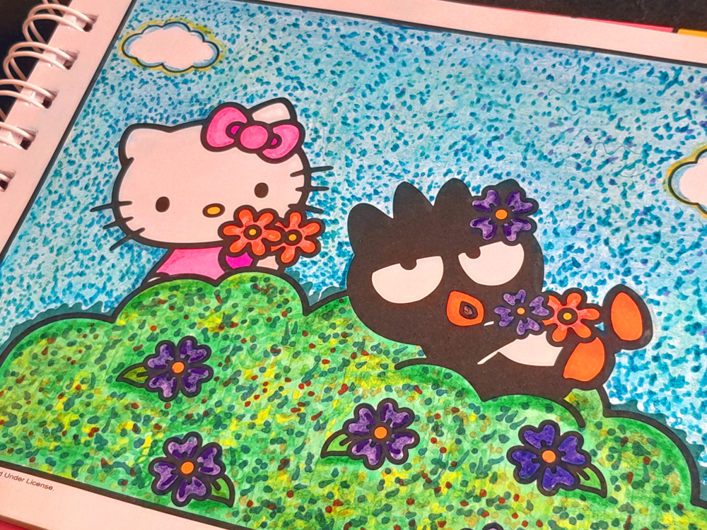 Página de livro de colorir já pintada, mostrando personagens da Hello Kitty em um cenário ao ar livre, com céu azul pontilhado, grama verde e flores coloridas. A pintura é feita com cuidado, pontilhado e cores vibrantes.