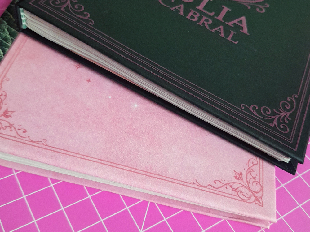 Detalhe de dois cadernos empilhados: um com capa verde-escura e acabamento elegante e outro com capa rosa suave e textura delicada. Ambos estão apoiados sobre uma base de corte rosa.