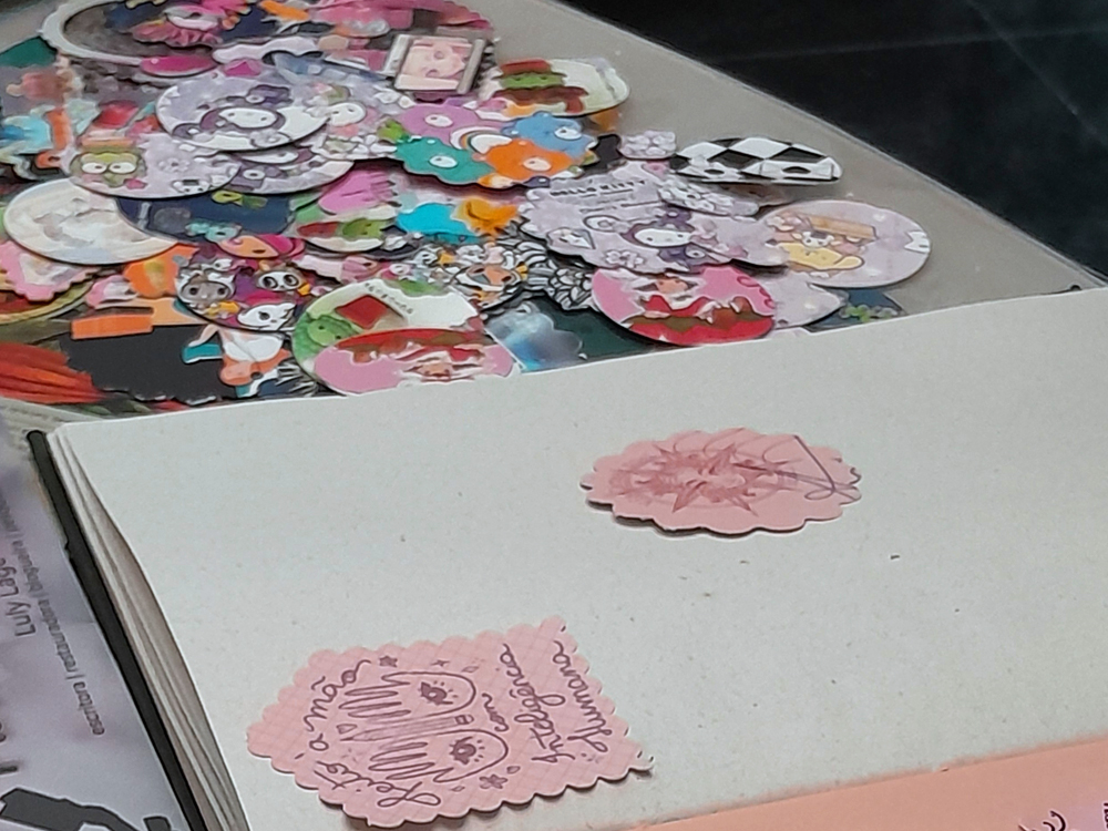 Mesa com um caderno aberto e vários adesivos coloridos espalhados ao fundo, em formatos variados e tons predominantemente rosa. Dois adesivos já estão colados na página.