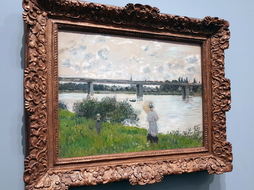 Pintura mostrando duas figuras humanas à beira de um rio, com vegetação verde em primeiro plano e uma ponte ao fundo. A cena é iluminada por luz natural suave, com pinceladas rápidas e cores claras, típica da produção de Monet na década de 1870–1880. A pintura está em uma moldura dourada trabalhada.