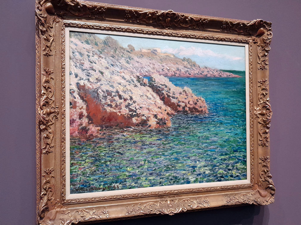 Pintura de Claude Monet retratando uma paisagem costeira com falésias rochosas em tons rosados e o mar azul-esverdeado em movimento. A superfície da água é construída com pinceladas densas e vibrantes. A obra, em moldura dourada ornamentada, possivelmente na região da Normandia.