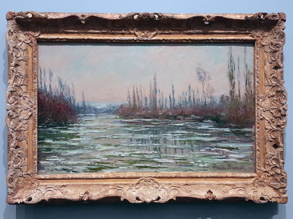Pintura de Claude Monet representando um curso d’água raso em paisagem aberta, com vegetação baixa e árvores esguias ao fundo, algumas ainda sem folhas. A água reflete tons de verde, cinza e rosado do céu, criando uma atmosfera fria e silenciosa. A obra está em moldura dourada ricamente entalhada, exposta em parede azul-clara no MASP. Assinatura do artista é visível no canto inferior direito.