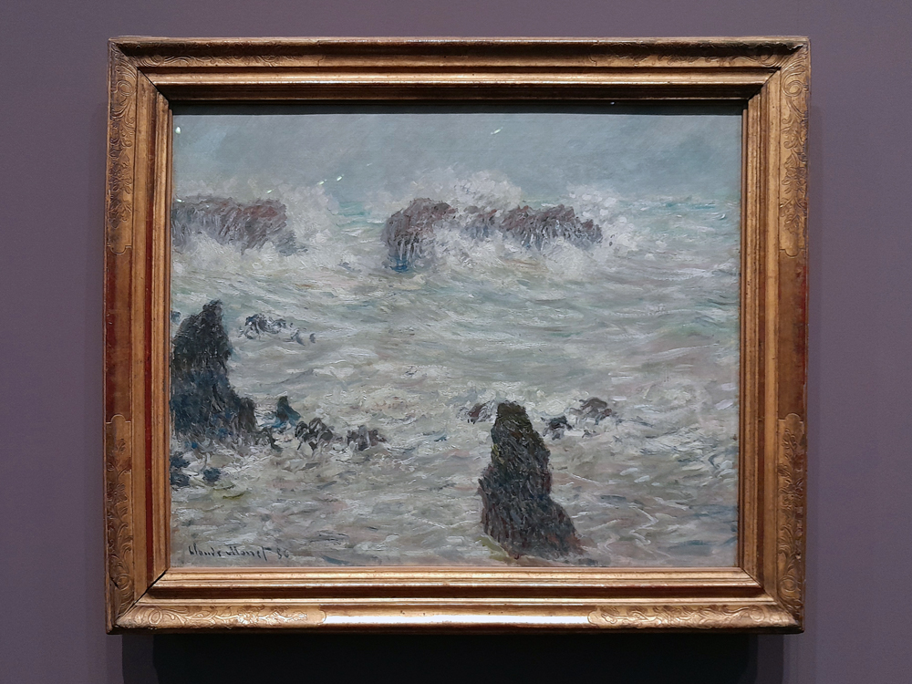 Pintura impressionista de Claude Monet representando um curso d’água raso com vegetação aquática e arbustos nas margens. A água reflete tons esverdeados e rosados do céu ao entardecer, enquanto árvores sem folhas surgem ao fundo, criando uma atmosfera silenciosa e úmida. A obra está emoldurada por uma moldura dourada ornamentada, exposta em parede azul.
