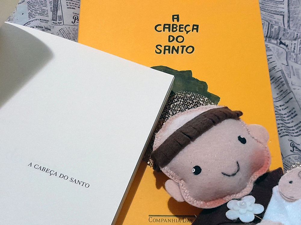 Livro aberto na folha de rosto, com o título impresso em tipografia simples, ao lado do boneco de santo em feltro, ambos sobre a capa amarela.
