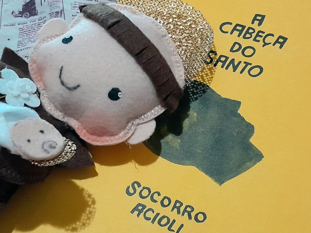 Detalhe do boneco de santo Antônio em feltro apoiado sobre a capa do livro A Cabeça do Santo, destacando o rosto bordado e a textura dos materiais.