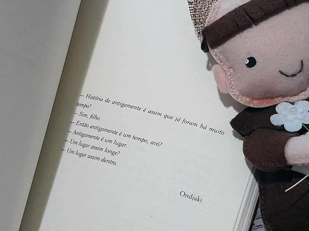 Livro aberto em página de texto que traz o diálogo: — História de antigamente é assim que já foram há muito tempo? — Sim, filho. — Então antigamente é um tempo, avó? — Antigamente é um lugar. — Um lugar assim longe? — Um lugar assim dentro., atribuída a Ondjaki.