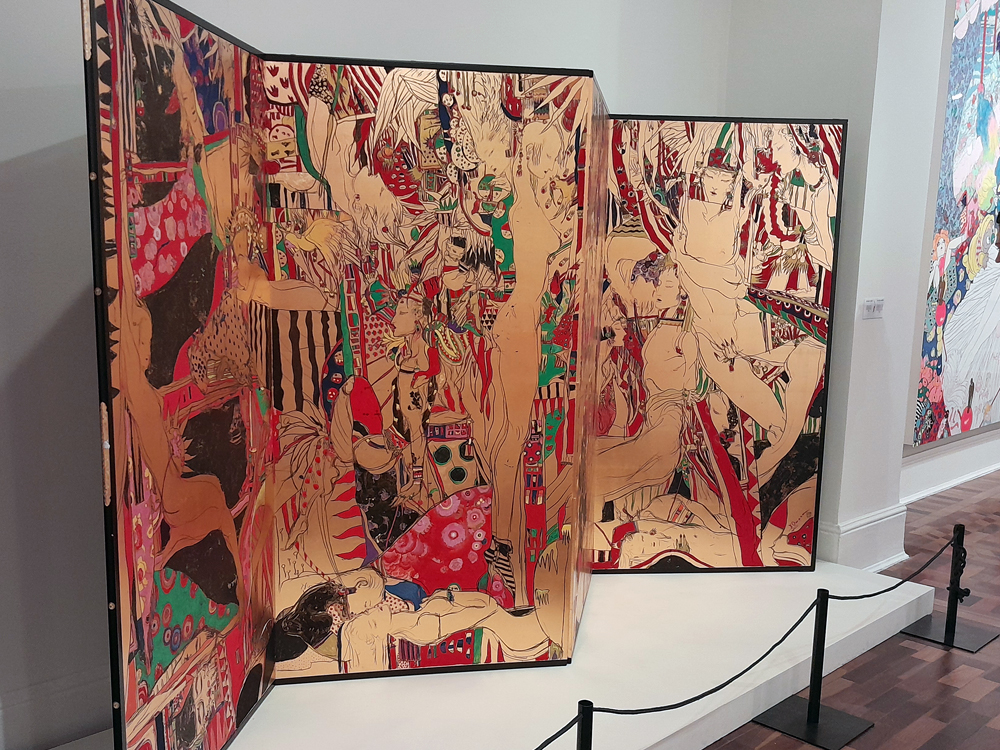 Painel dobrável de grandes dimensões com ilustrações de Yoshitaka Amano, repleto de figuras humanas estilizadas, corpos entrelaçados e padrões ornamentais em tons de vermelho, bege e preto, instalado no espaço expositivo do CCBB BH.