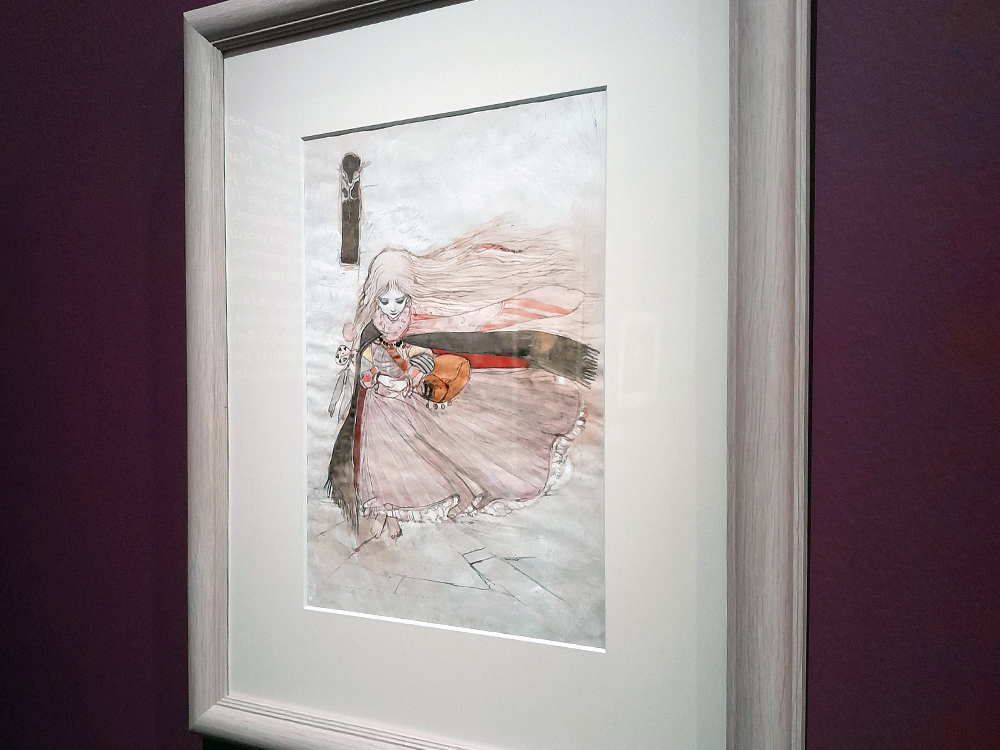 Obra de Yoshitaka Amano emoldurada, mostrando figura feminina de vestido esvoaçante caminhando em cenário melancólico, com linhas suaves, tons claros e atmosfera poética, exposta sobre parede em tom roxo no CCBB BH.