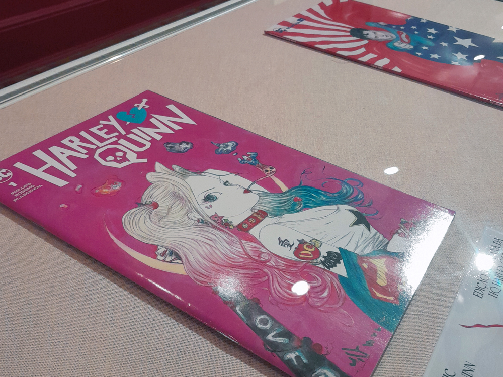 Revista em quadrinhos de Harley Quinn, em exibição sob vitrine, com capa em tons de rosa e azul.