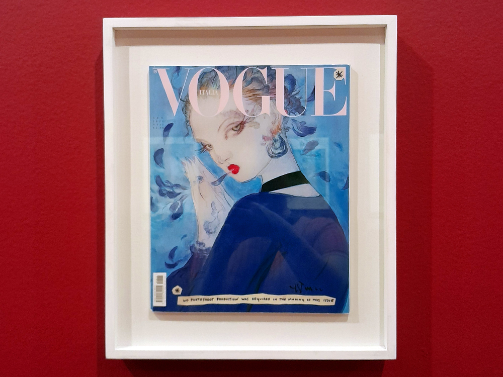 Capa da revista Vogue Itália, com figura feminina estilizada em tons de azul e vermelho, emoldurada e exibida na exposição.