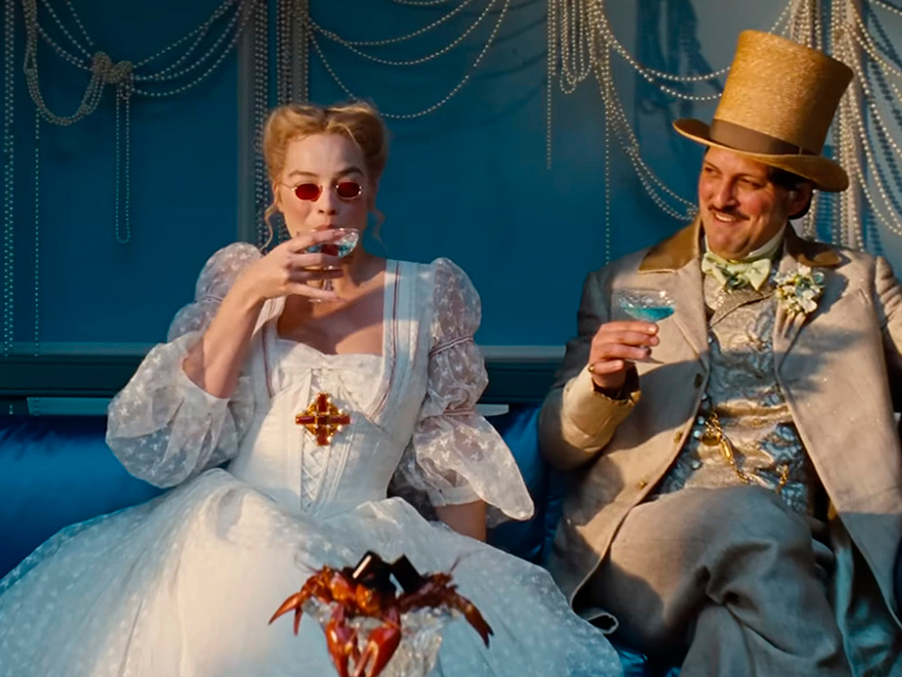Cena do filme onde Catherine sentada em um sofá azul, vestindo um vestido branco de mangas bufantes e óculos de lentes vermelhas, enquanto toma uma bebida azul em taça. Ao seu lado, um homem elegante com cartola também segura um drink azul. O cenário inclui cortinas de pérolas e iluminação em tons azulados, criando uma atmosfera visualmente luxuosa e excêntrica.