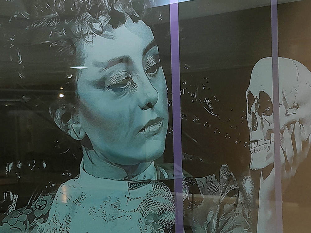Close de uma projeção ou painel com imagem em tons azulados de uma atriz clássica do cinema observando um crânio humano, evocando a cena de Hamlet e criando um contraste dramático entre o rosto da personagem e a caveira iluminada.