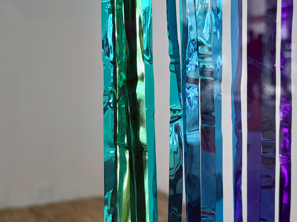 Detalhe de tiras verticais metálicas e refletivas em diferentes tons de azul, verde e roxo, penduradas lado a lado como uma cortina brilhante que distorce reflexos e cria efeitos de luz na superfície.