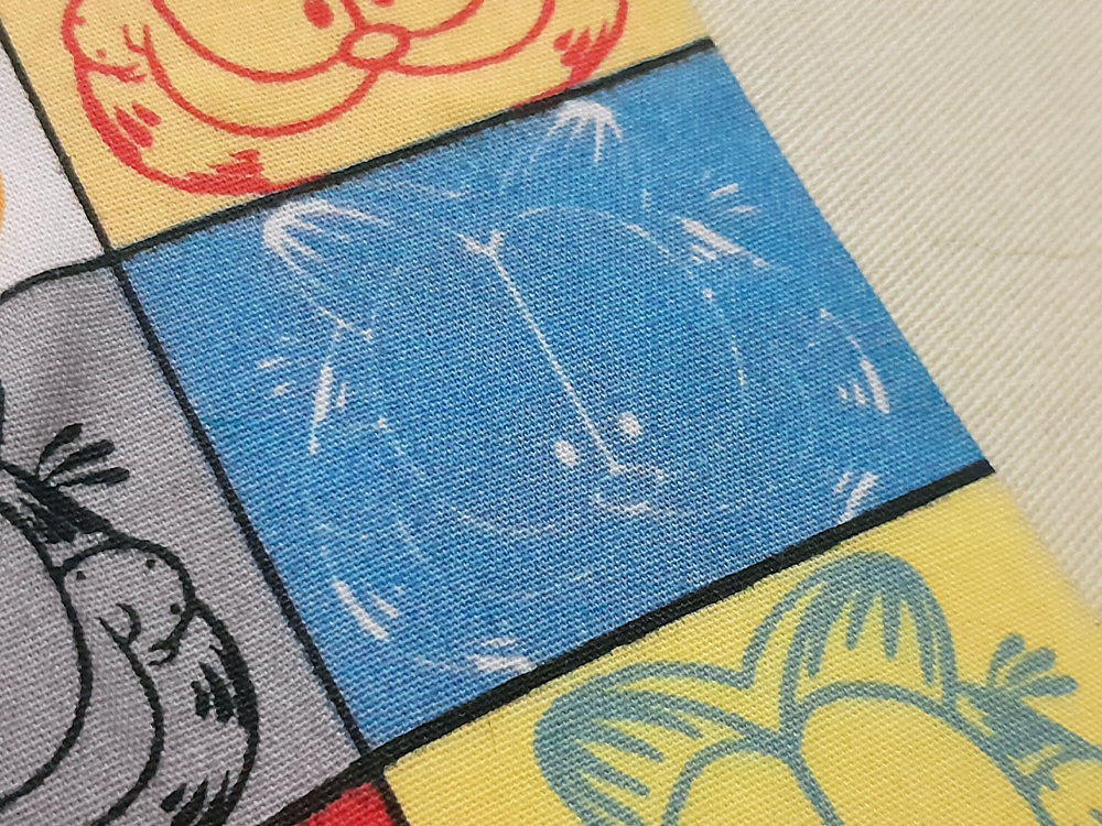 Close de tecido estampado dividido em quadrantes coloridos com o rosto do gato Garfield, incluindo um quadro azul com desenho branco, cercado por outras ilustrações e padrões gráficos em amarelo, vermelho e preto.