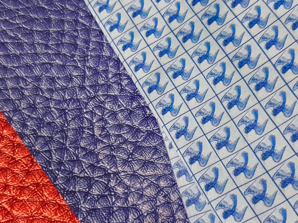 Composição de texturas e materiais sobrepostos: superfície sintética azul escura, simulando couro, tecido quadriculado com repetição de pequenos pássaros azuis impressos e um recorte de material vermelho metálico brilhante.