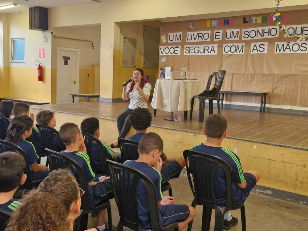 Mulher com microfone falando para alunos sentados em frente ao palco durante evento literário na escola Barão de Macaúbas, com decoração temática e frase sobre leitura ao fundo.