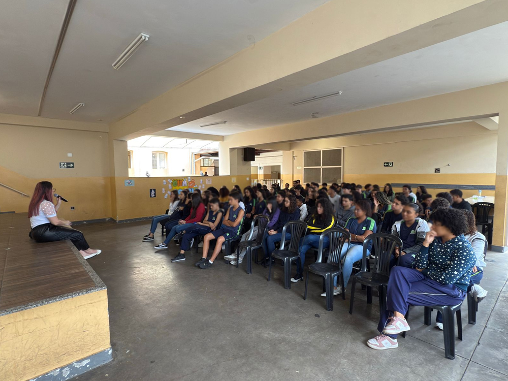 Vista lateral de auditório cda escola Barão de Macaúbas om estudantes atentos enquanto uma mulher fala ao microfone no palco durante atividade escolares.