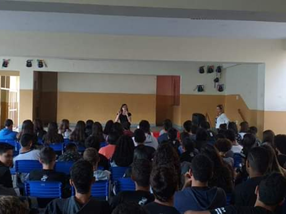 Auditório escolar com grande grupo de estudantes assistindo a uma apresentação no palco, onde duas pessoas estão à frente conduzindo a atividade.